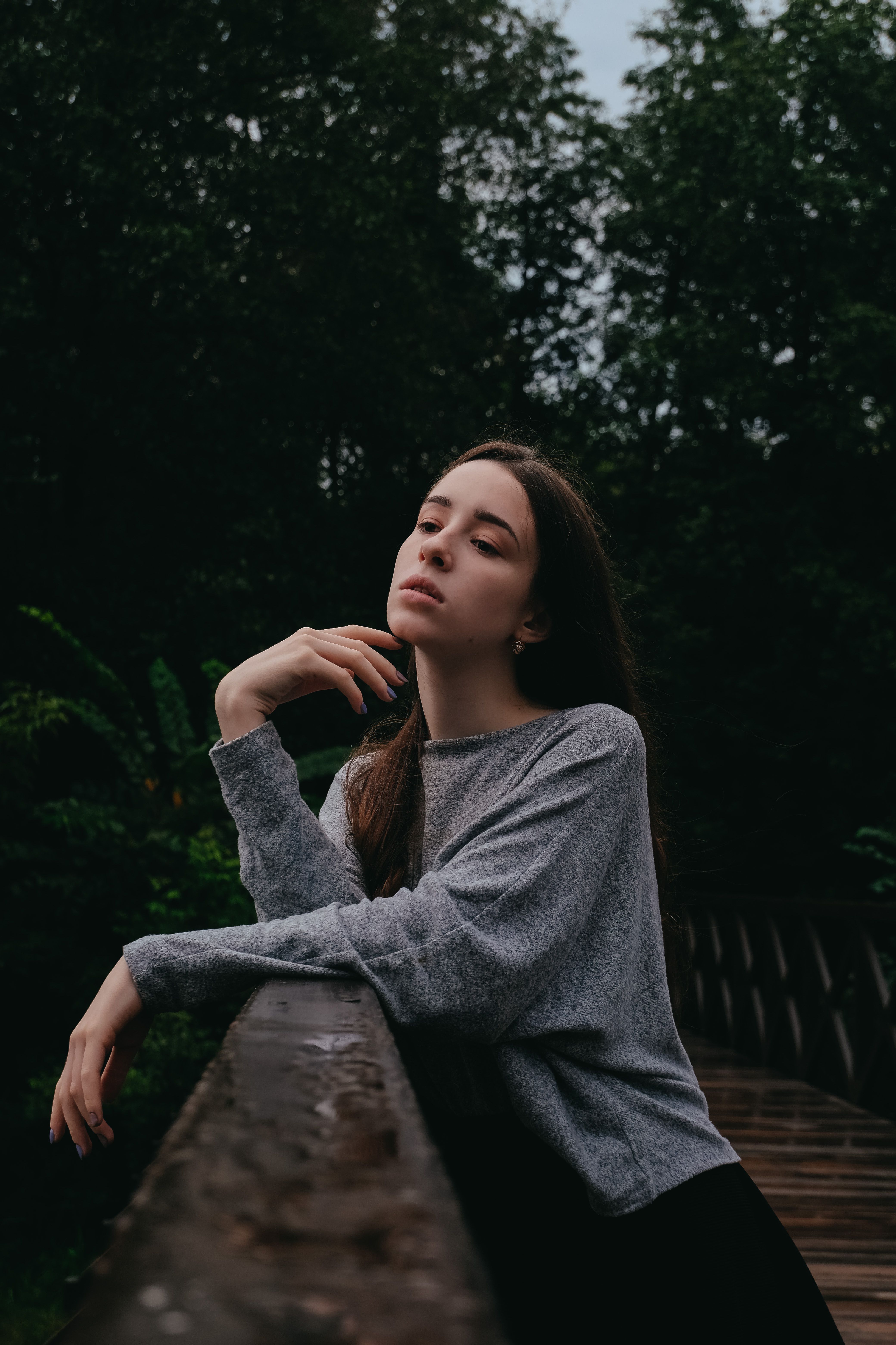 一位棕色长发、面容姣好的女子，站在森林中的绿色桥梁上，倚着栏杆若有所思地向下望去