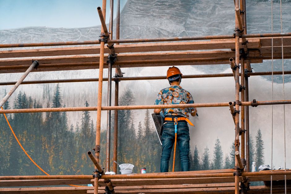 Worker on scaffolding painting a large forest landscape mural.