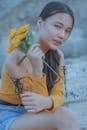 Young female looking at camera while holding bouquet of artificial flowers and resting on rough rocky surface on blurred background of nature