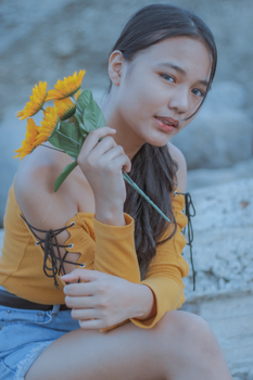 Young female looking at camera while holding bouquet of artificial flowers and resting on rough rocky surface on blurred background of nature