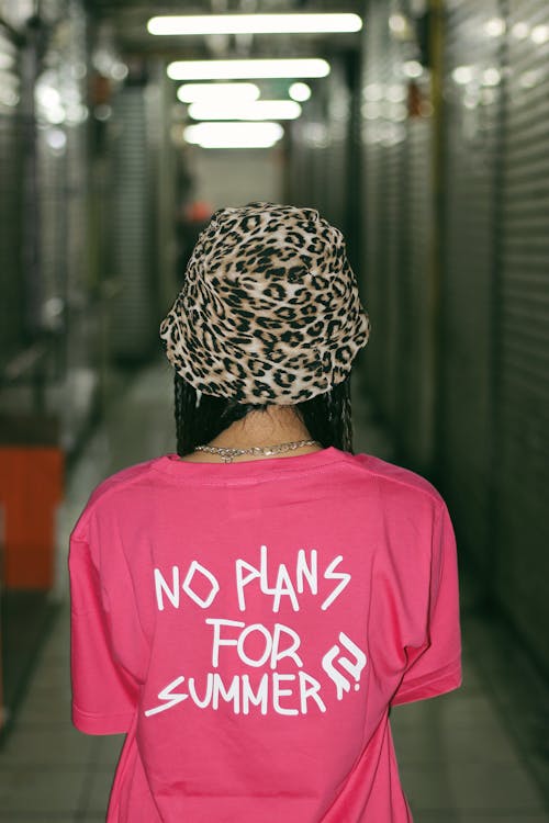 Back view of unrecognizable female teenager in stylish hat and t shirt with No Plans For Summer inscription standing in corridor of modern building