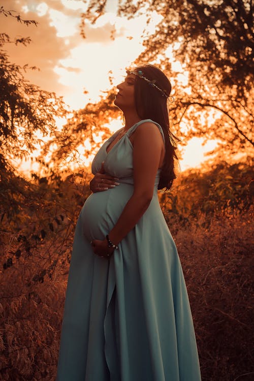 Side view of pregnant female in long dress with wreath on head standing on grassy lawn near branches with hand on belly