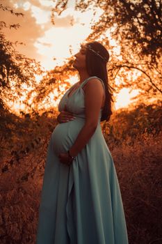 Side view of pregnant female in long dress with wreath on head standing on grassy lawn near branches with hand on belly - Fotografer Sidoarjo
