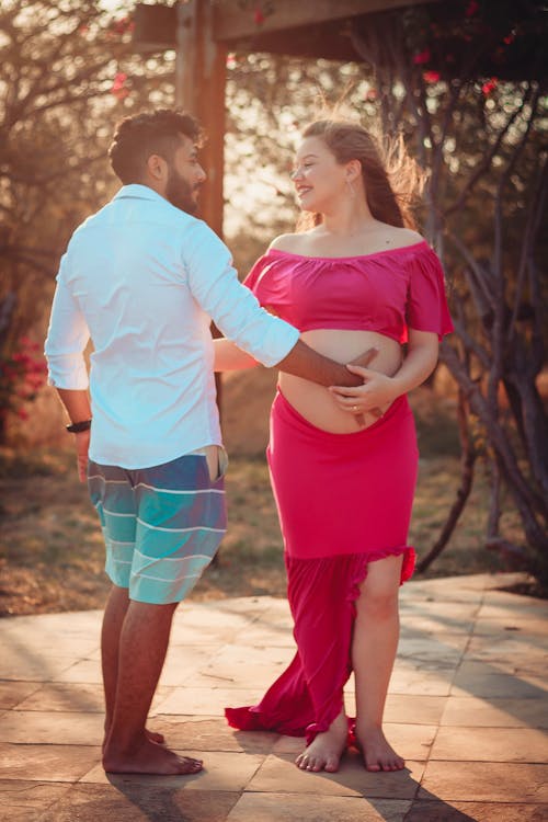 Full body of pregnant barefoot female in top standing near ethnic male with hand on bare belly while looking at each other