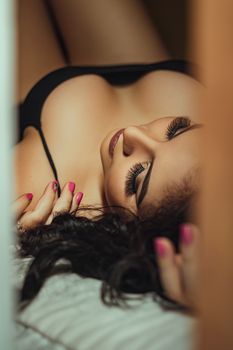 A seductive woman with dramatic makeup and lashes lies in black lingerie.