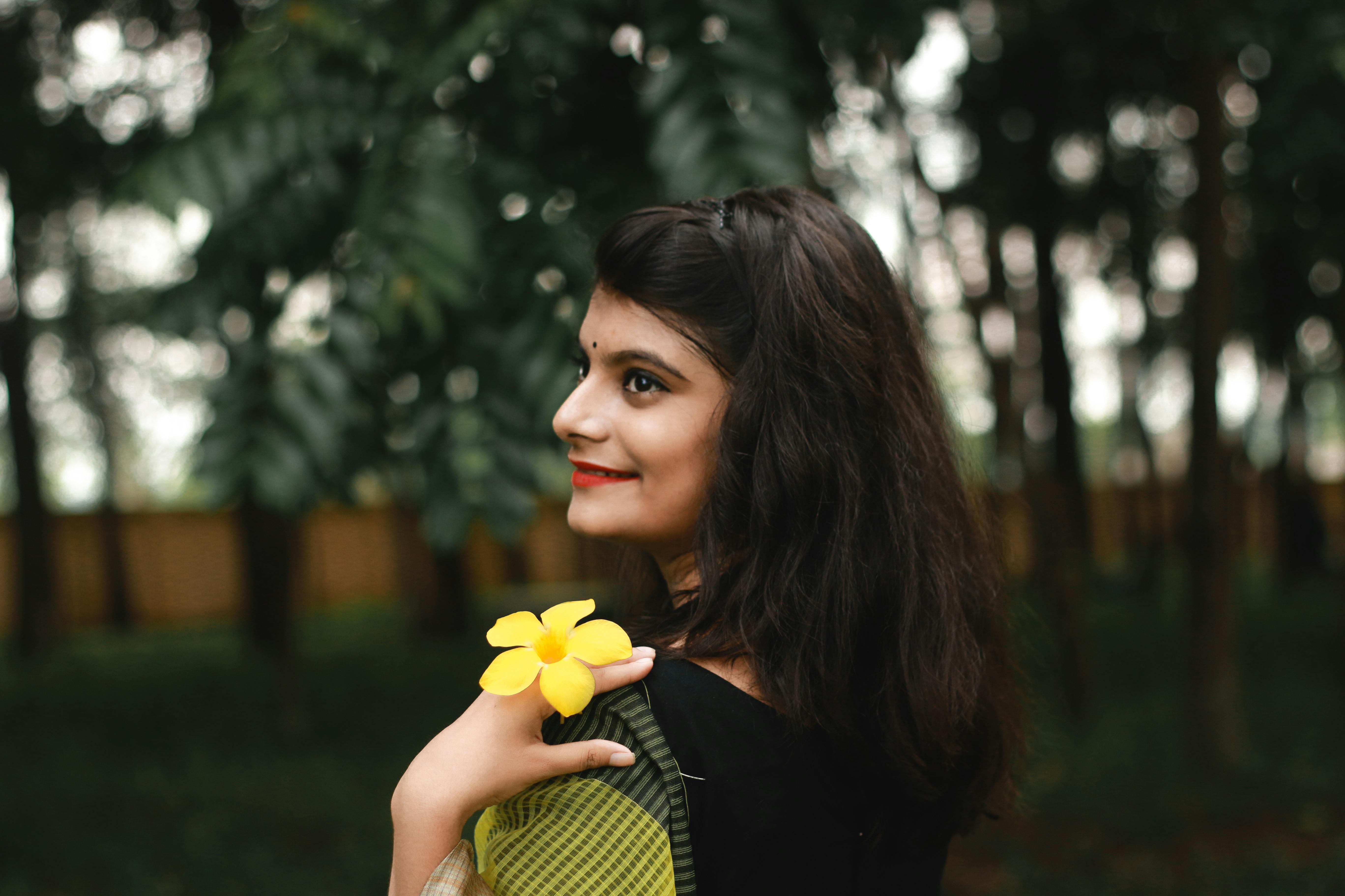 Free Side view of young cheerful ethnic tfemale with yellow flower looking away on blurred background of green trees Stock Photo