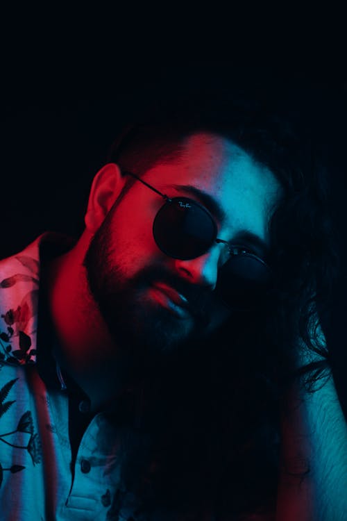 Confident bearded brutal guy in stylish sunglasses under bright light in dark room