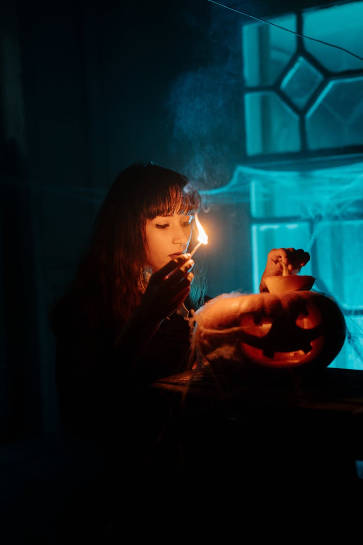 Woman In Black Long Sleeves Holding Lighted Match Stick While Looking At The Jack O Lantern