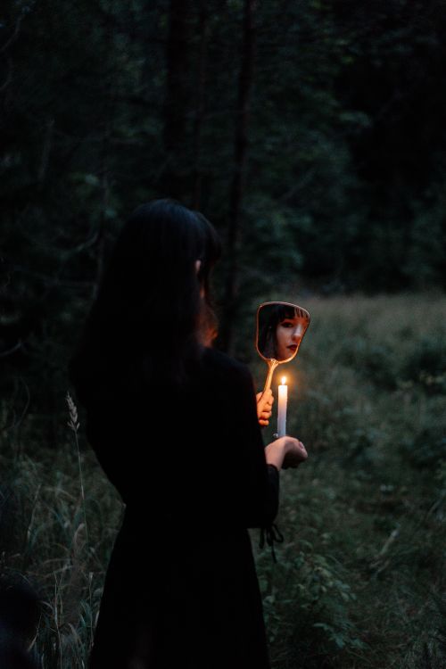 Free A woman holds a candle and mirror in a dark forest, reflecting a mystical, eerie mood. Stock Photo
