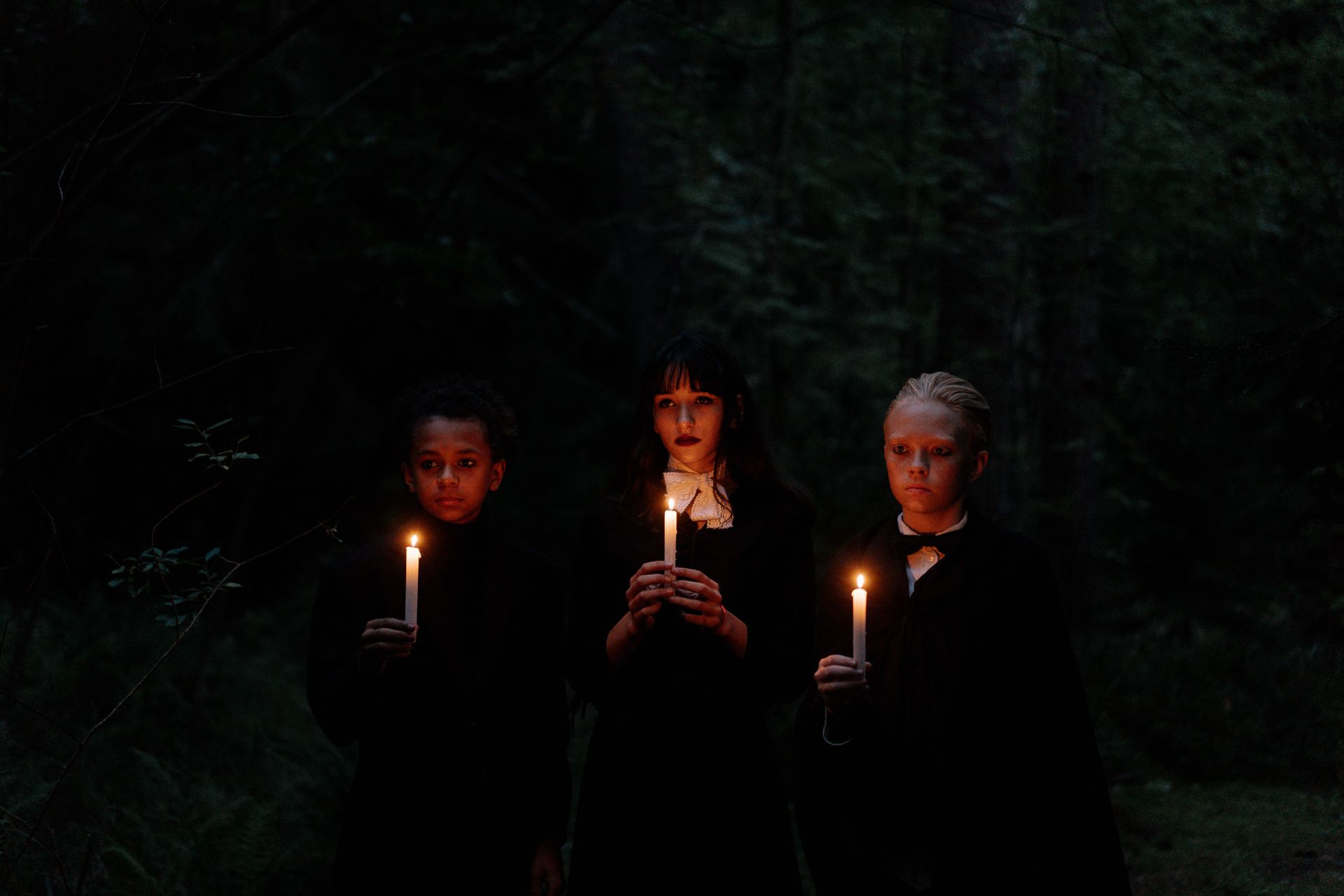 Three teenagers in dark clothing holding candles, creating an eerie and spooky atmosphere in a forest at night.