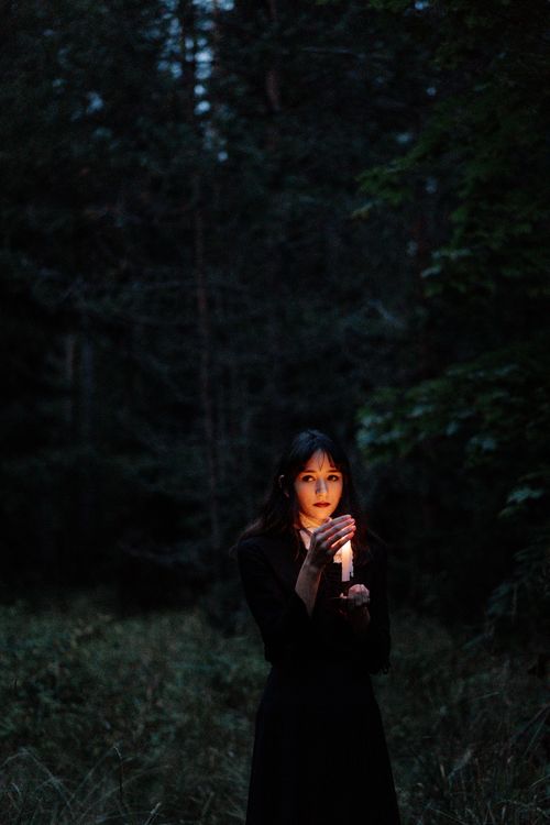 Free A young woman stands with a candle in a dark, eerie forest setting. Stock Photo