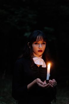 一位身着哥特式风格的年轻女子手持蜡烛，在黑暗的森林中营造出一种怪诞而神秘的氛围