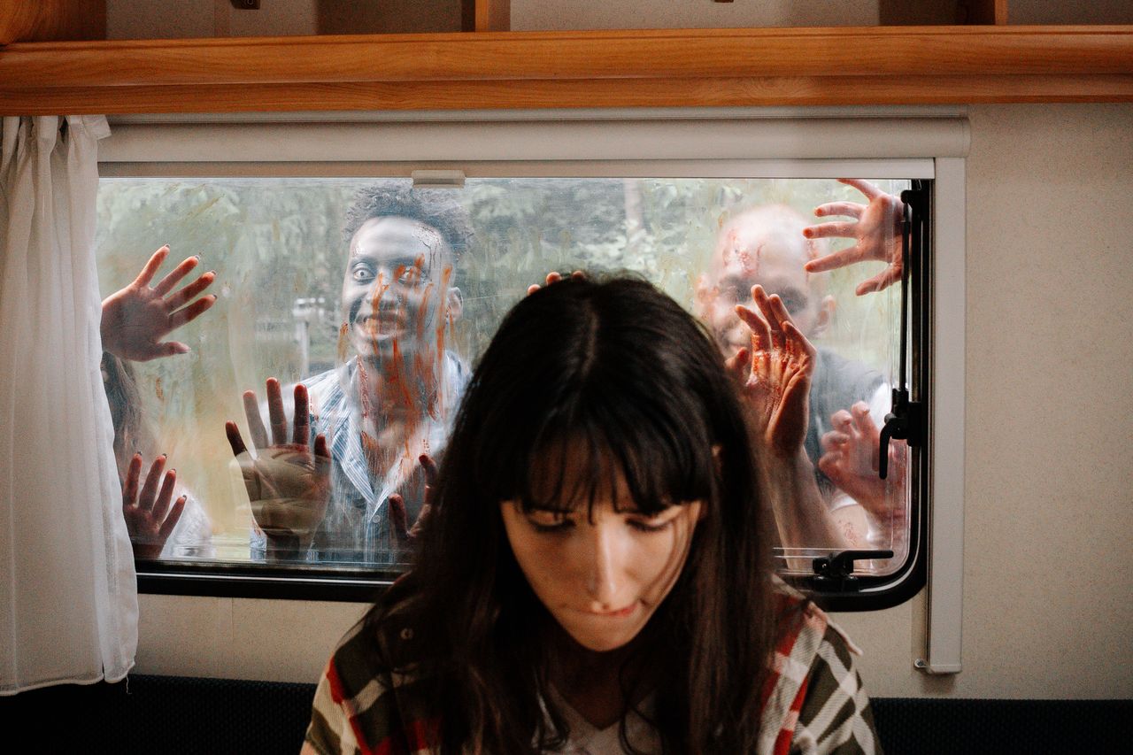 a woman sits unaware while terrifying zombies threaten through the win