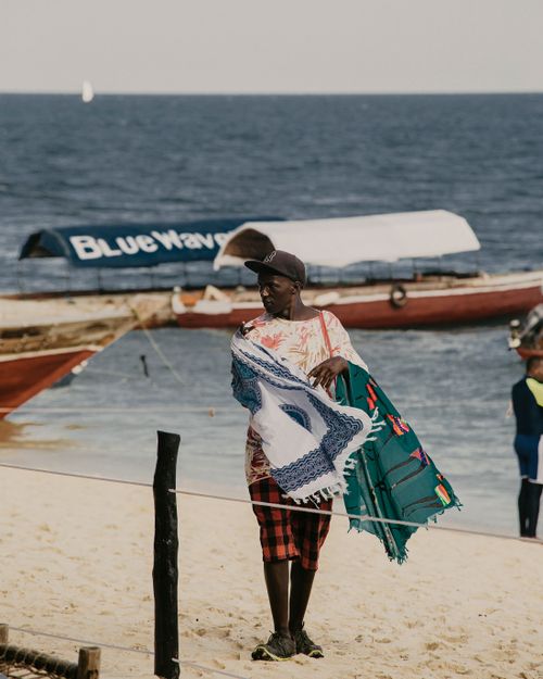 Free African vendor selling colorful scarves on a serene beach. Stock Photo