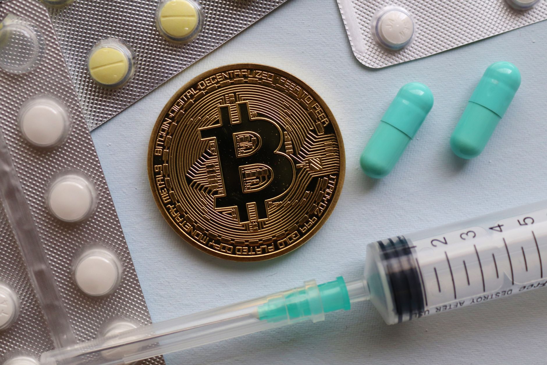 A close-up of a bitcoin, pills, and a syringe, highlighting intersections of finance and healthcare.