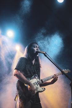 Indie musician performing live with guitar amidst vibrant stage lights and smoke.