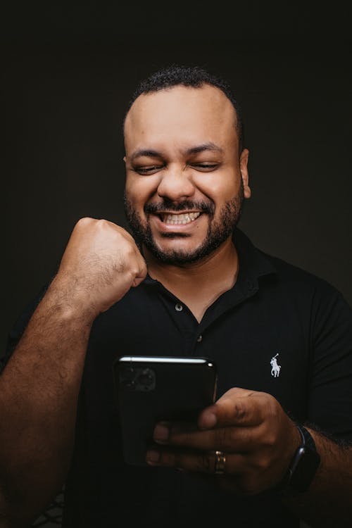 Handsome bearded male with black short hair and classic t shirt holding smartphone in hand and smiling happily