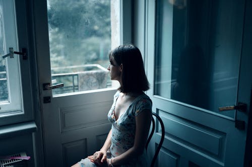 Young female in casual dress sitting on chair near white door and looking at window
