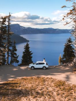 From above of anonymous traveler standing near modern white SUV car and admiring spectacular view of mountain lake against cloudy blue sky