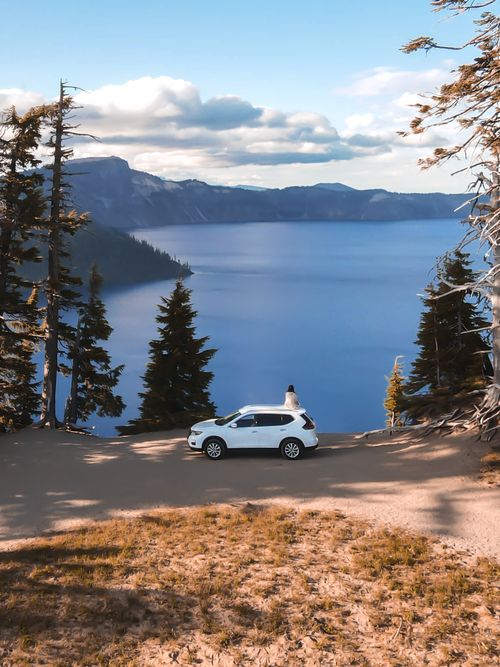Free From above of anonymous traveler standing near modern white SUV car and admiring spectacular view of mountain lake against cloudy blue sky Stock Photo