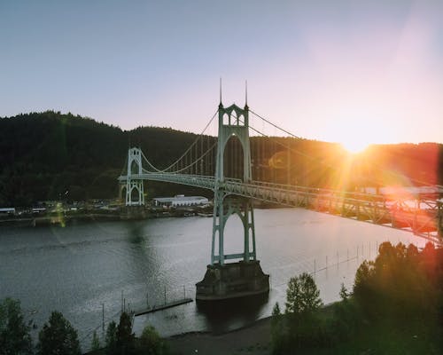 Magnificent scenery of aged St Johns Bridge crossing rippling river surrounded by hills with lush green woods against cloudless sunset sky in Portland