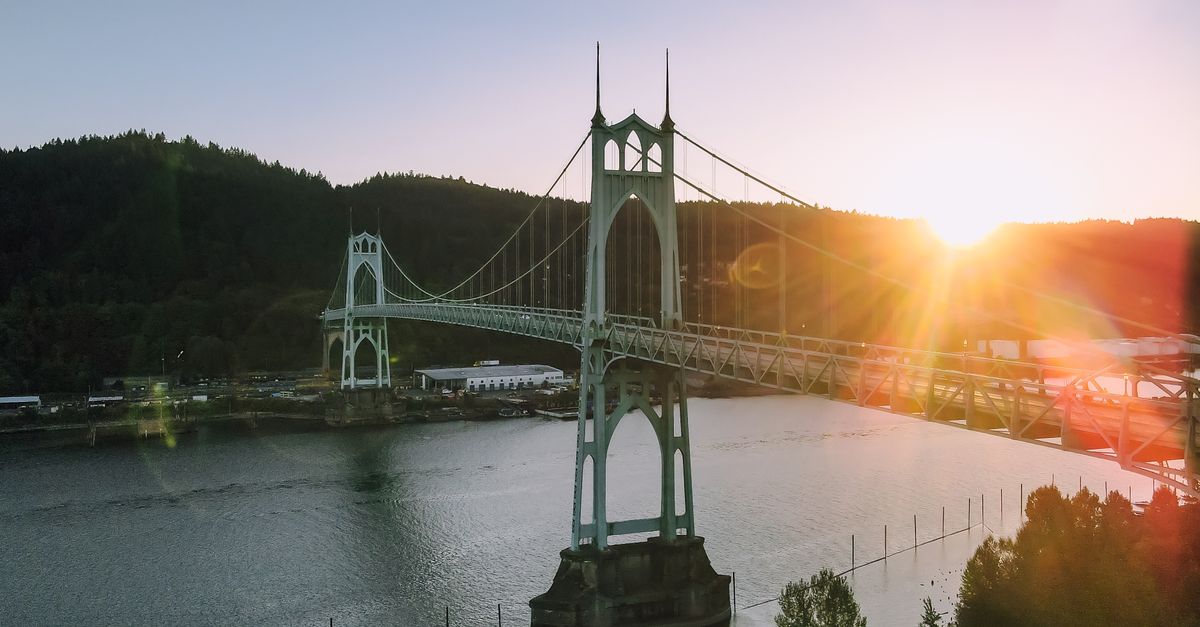 Magnificent scenery of aged St Johns Bridge crossing rippling river surrounded by hills with lush green woods against cloudless sunset sky in Portland