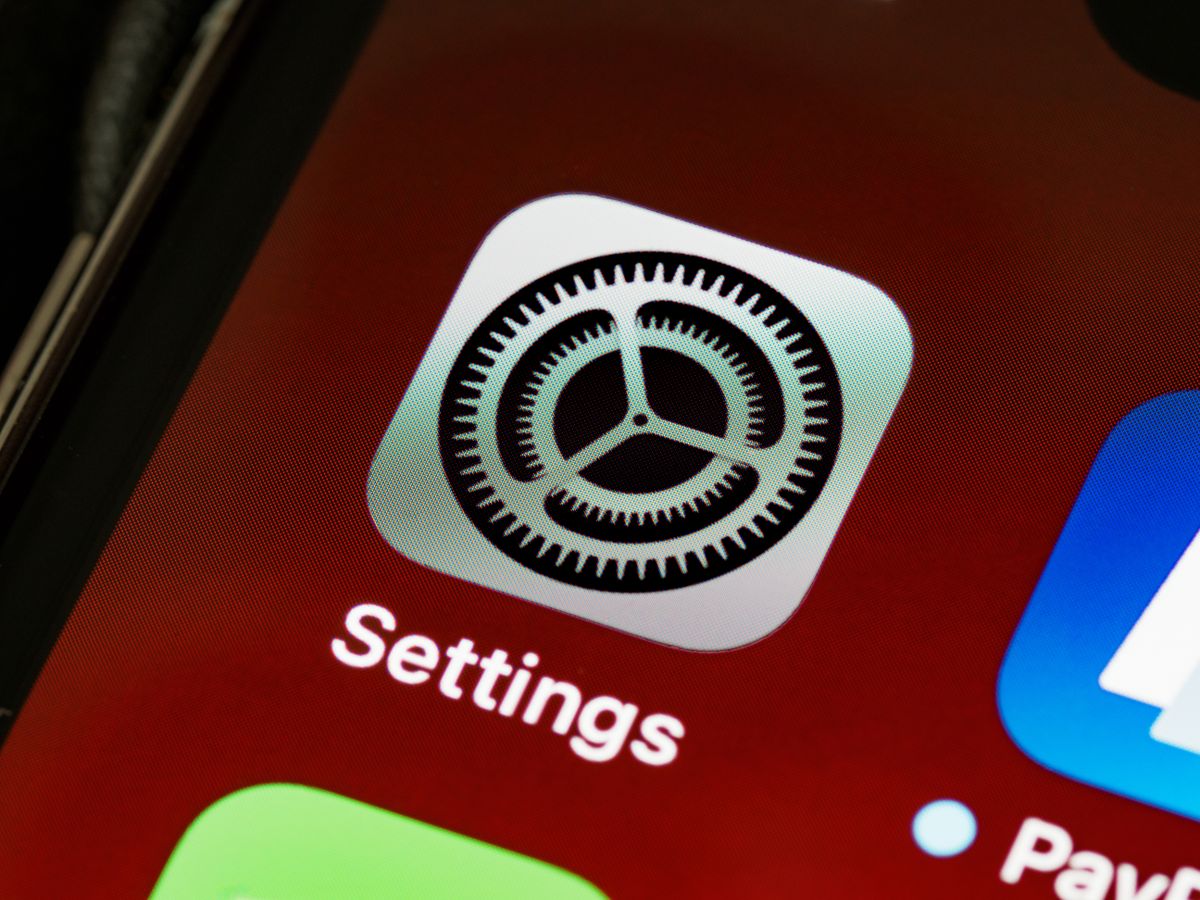 A close up of an iPhone home screen filled by the grey Settings gear icon on a deep red background, with the corner of a blue app and a green app visible at the edges