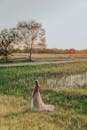 From above full body of distant unrecognizable female in long dress with hem strolling in grassy meadow with tall trees in sunlight