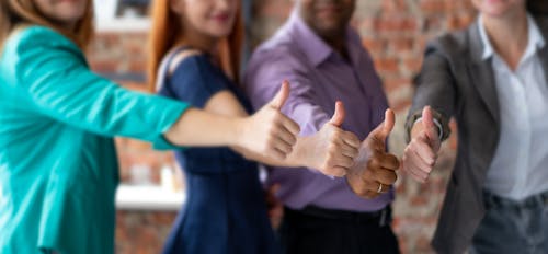 Unrecognizable cheerful colleagues in formal clothes showing thumbs up gesture while standing in modern office during teamwork on blurred background