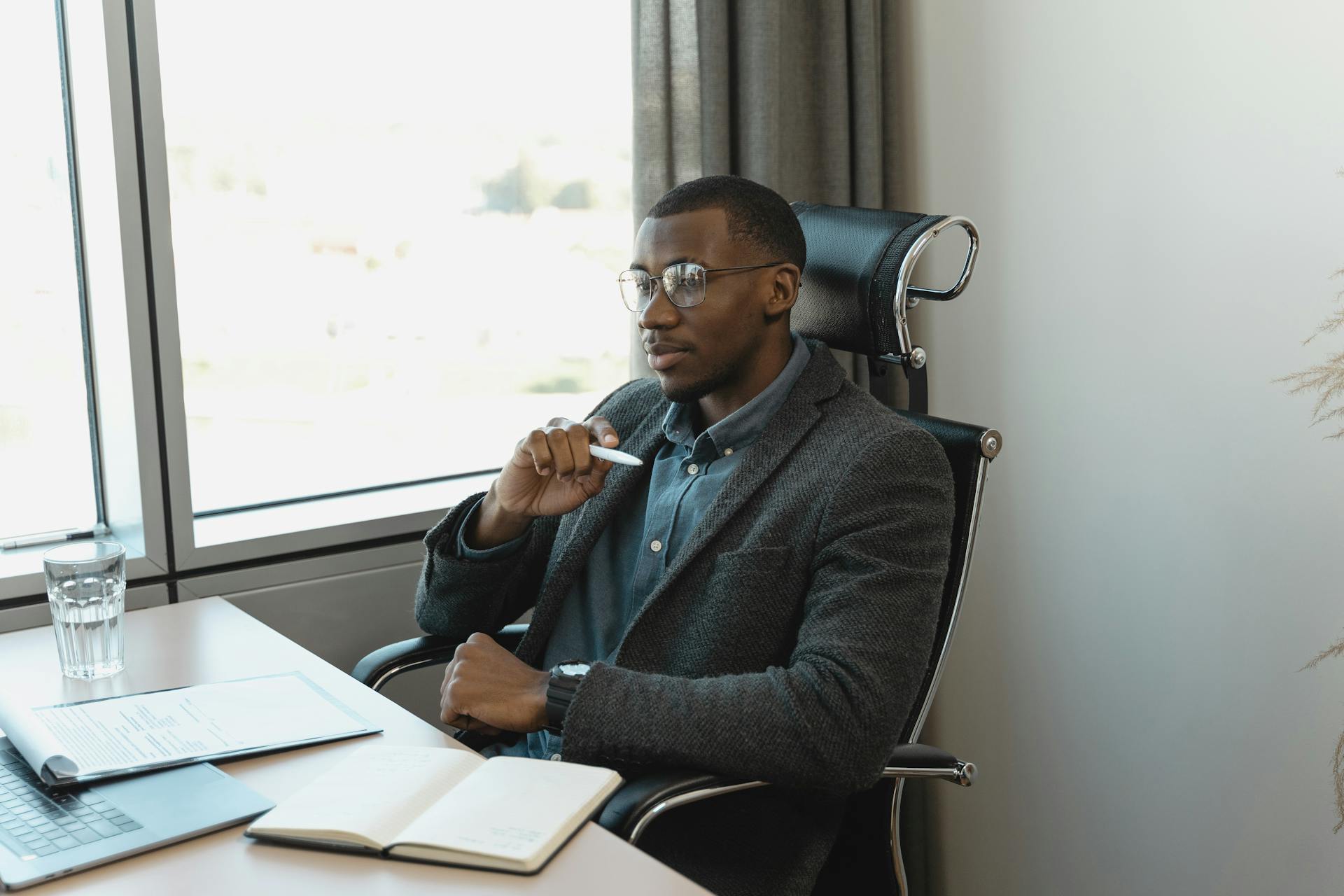 Pensive businessman sitting at a desk, thinking with a pen in hand in a modern office setting.
