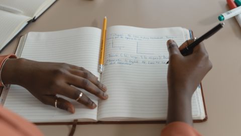 Focused close-up of hands writing in a notebook with a pen and pencil on a table.