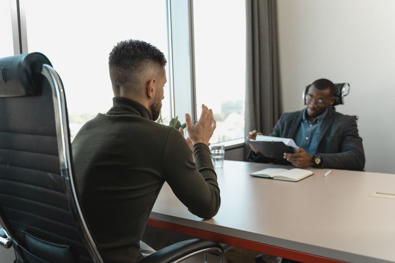A candidate speaking across the table while an interviewer reviews notes in a bright meeting room