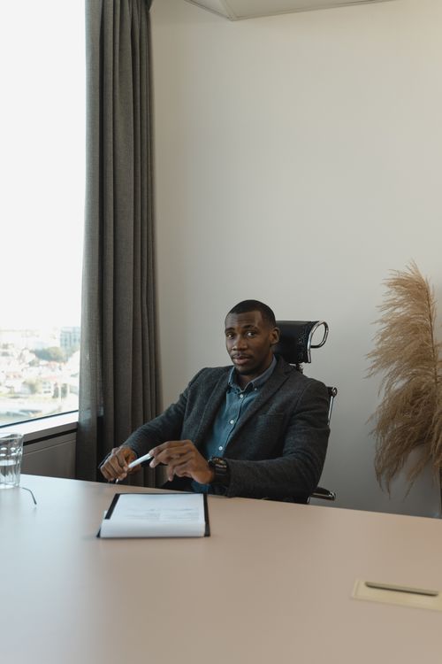 Free Confident businessman holding a pen and clipboard, seated at a desk in a modern office setting. Stock Photo