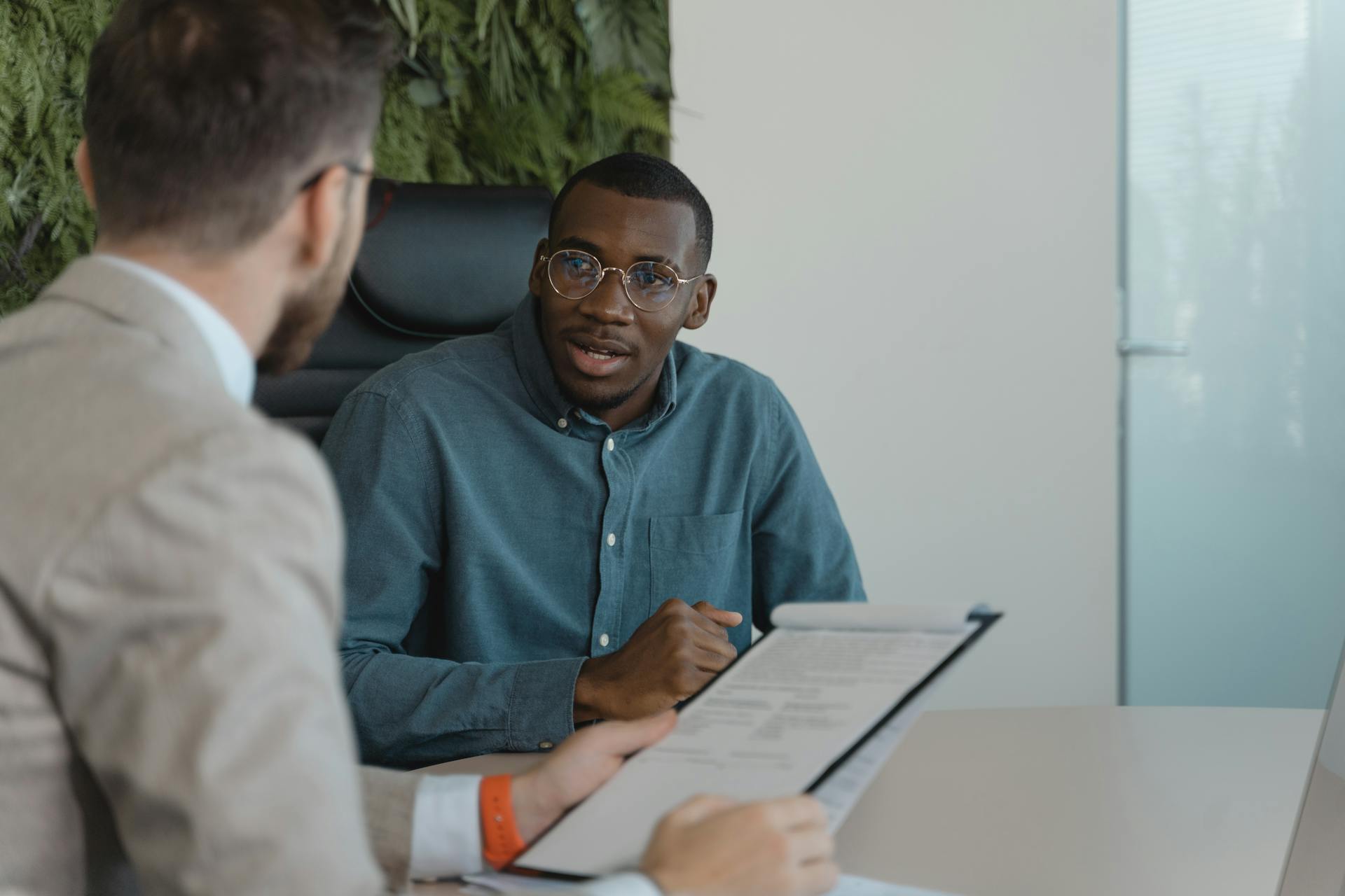 Two men engaged in a professional meeting at an office table with documents and a plant wall background.