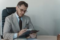 办公室里，一位穿着灰色西装、戴着眼镜的商人正在查看剪贴板上的文件