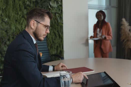 Focused businessman using tablet in modern office with green wall and colleague in background.