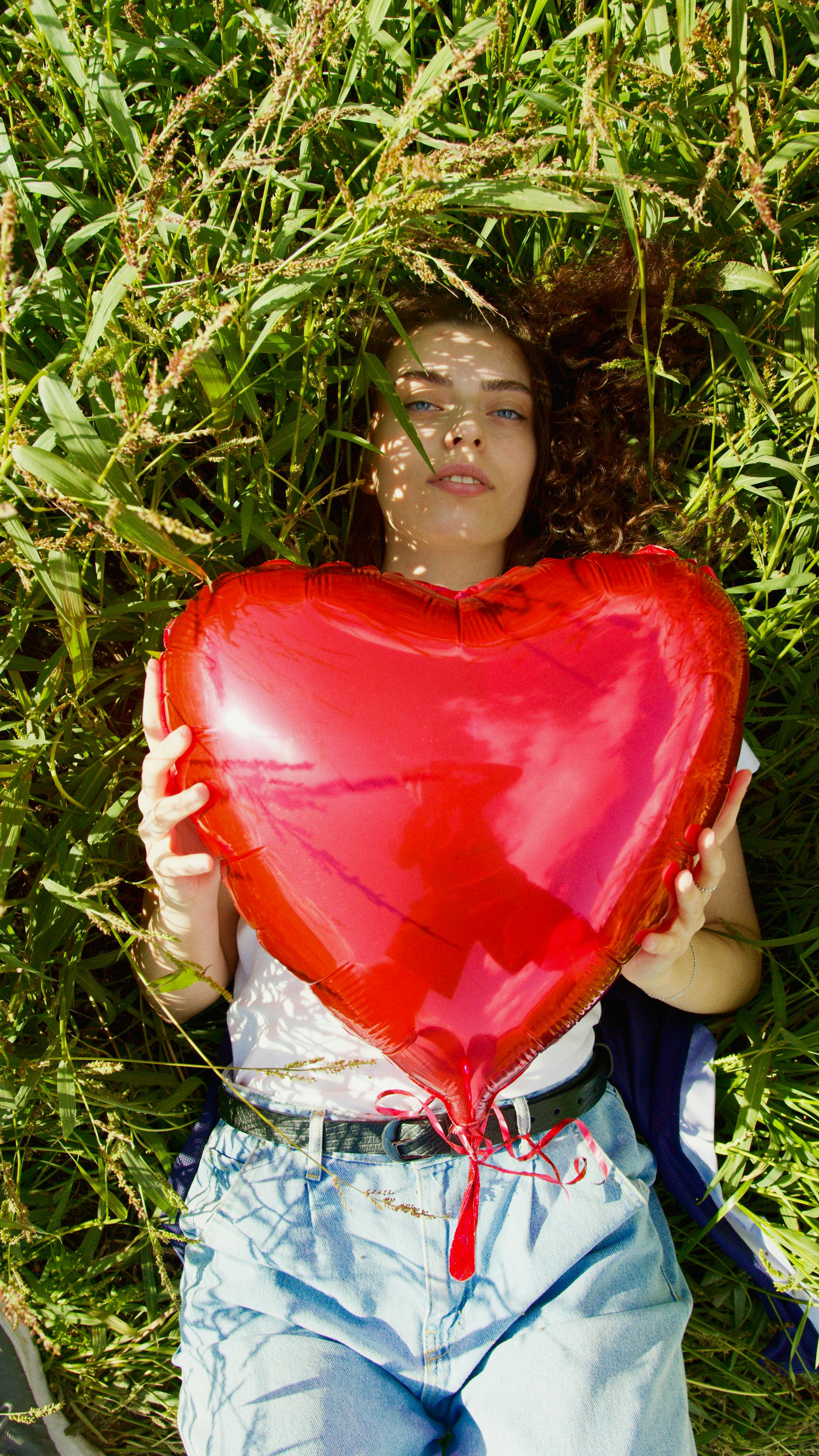 Young woman relaxing outdoors while holding a vibrant red heart-shaped balloon, evoking warmth and romance.