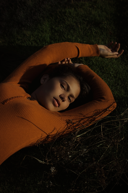 From above of tanned female looking at camera and resting in fresh green grass while stretching arms