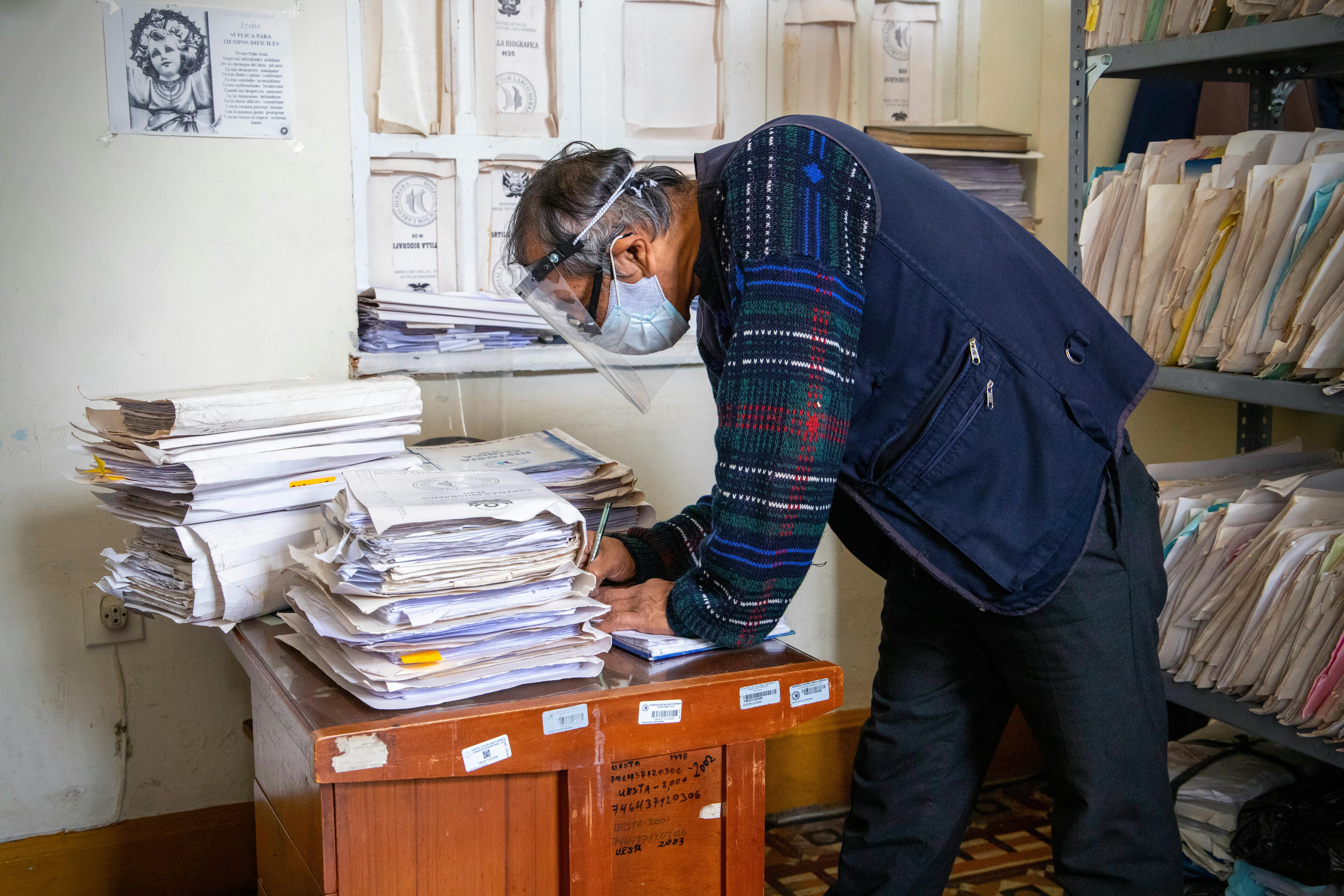 Elderly man with face shield sorts through piles of documents in an office setting.