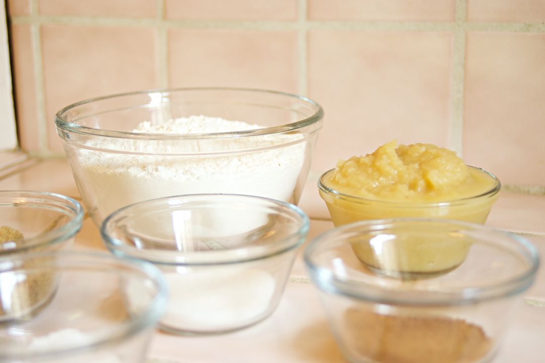 How to Price Your Baked Goods: The Complete Formula for Home Bakers in India (2026) 1 Glass bowls of flour, sugar, and other baking ingredients arranged on a kitchen counter for baking cost calculation.