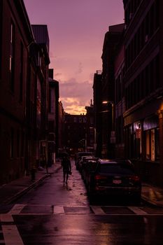 Vivid pink sky over a Boston street at dusk with silhouettes of buildings and a walker.