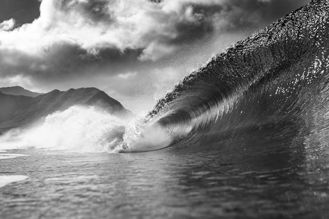 Black and white of foamy wave falling into ocean with splashes against cloudy sky in summer day in nature outside