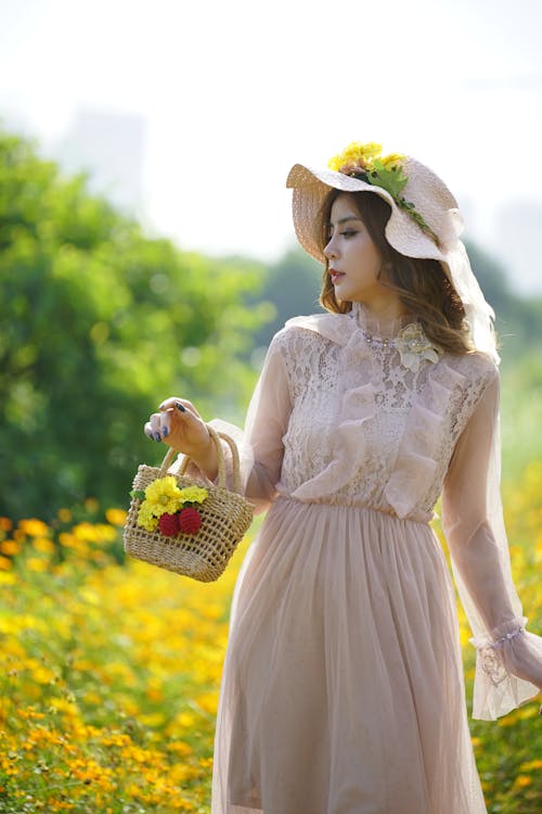 Free Stylish female in dress with wicker handbag standing in blooming field with yellow flowers on sunny summer day in countryside Stock Photo