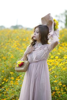 夏日，一位身着连衣裙、手提柳条包的恬静女子闭眼站在草地上，四周盛开着黄色的花朵