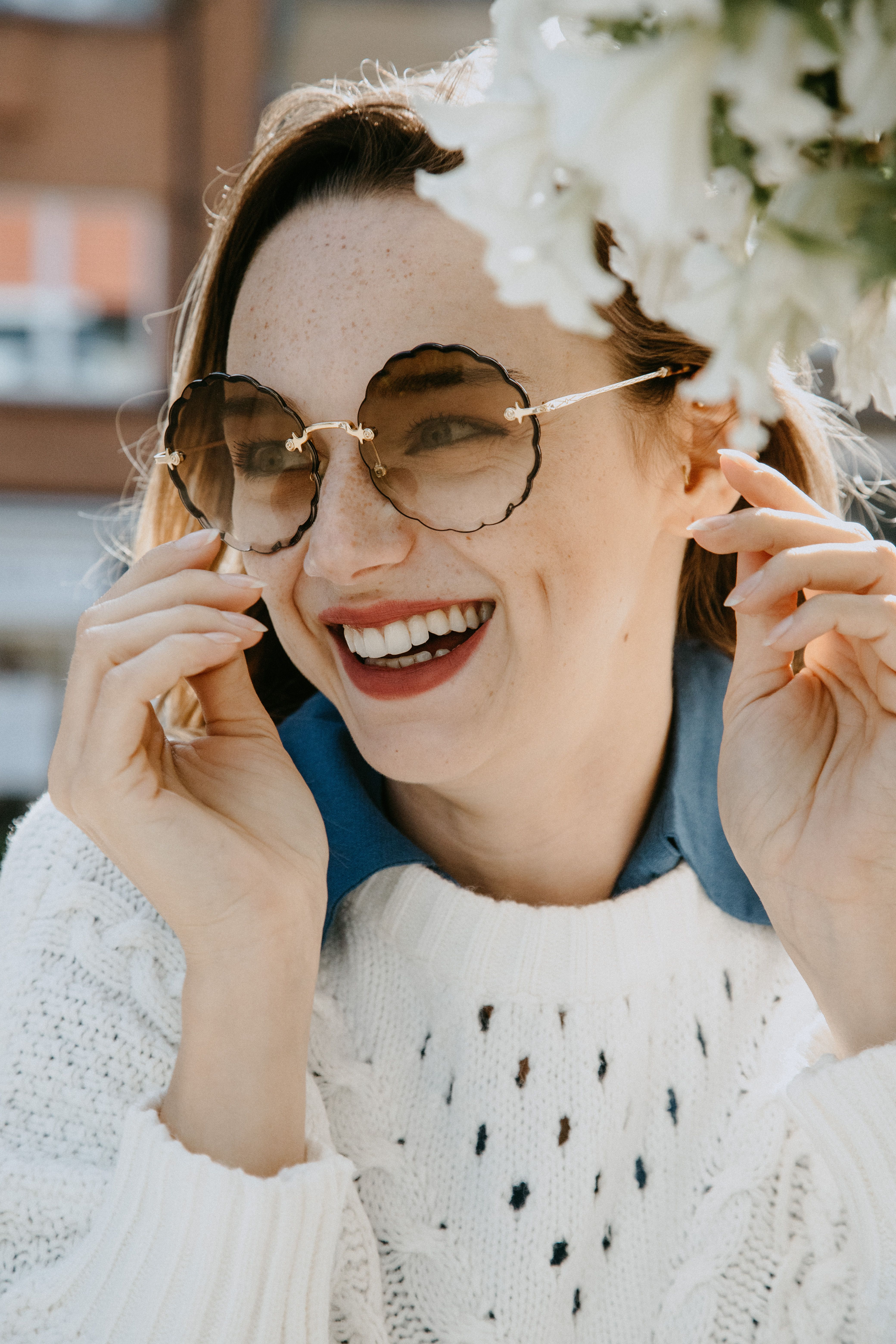 A cheerful woman with sunglasses laughs under the sun, showcasing her red lips and bright smile.