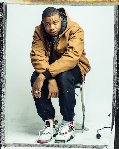 Full body calm young African American male in street style clothing and unlaced sneakers sitting on chair against white wall and looking at camera