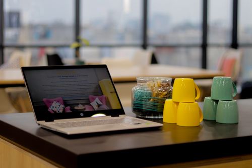 Free Sleek laptop on a desk with colorful stacked mugs in a bright office setting. Stock Photo