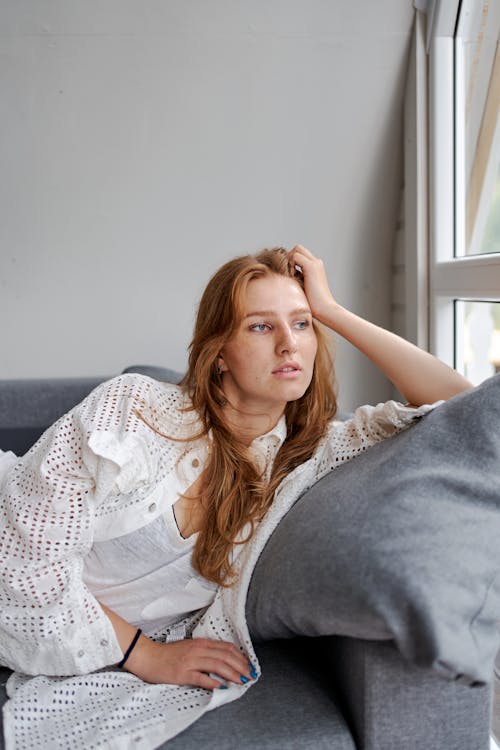 Young pondering female resting on cozy couch near window while looking away in house