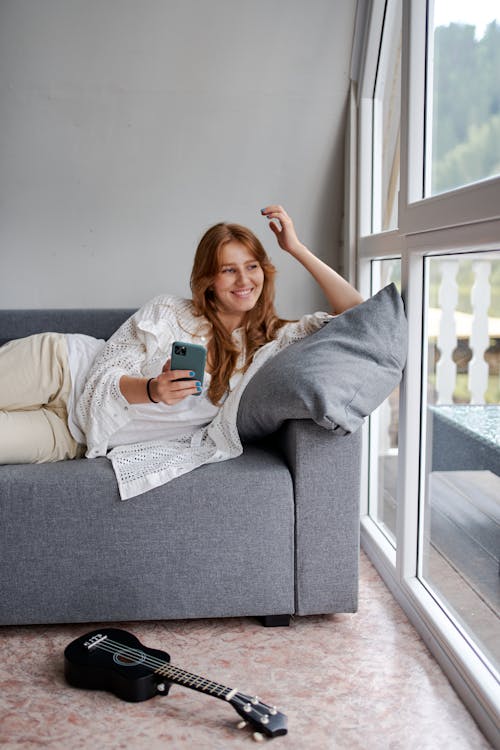 Free Young content female with cellphone resting on comfortable couch near ukulele on floor and glass wall while looking away Stock Photo