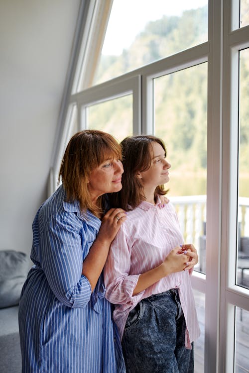 Side view of mother and daughter in casual clothes standing close together and dreaming while looking out big window